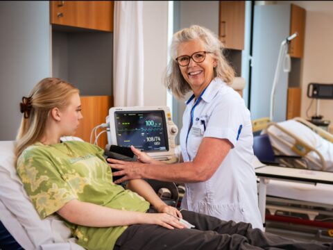 A nurse wearing glasses and a white uniform is smiling while measuring the blood pressure of a young woman lying on a hospital bed. The young woman is wearing a green tie-dye shirt and dark pants. A medical monitor displaying vital signs is visible behind them. The setting appears to be a hospital room with medical equipment and a bed in the background.